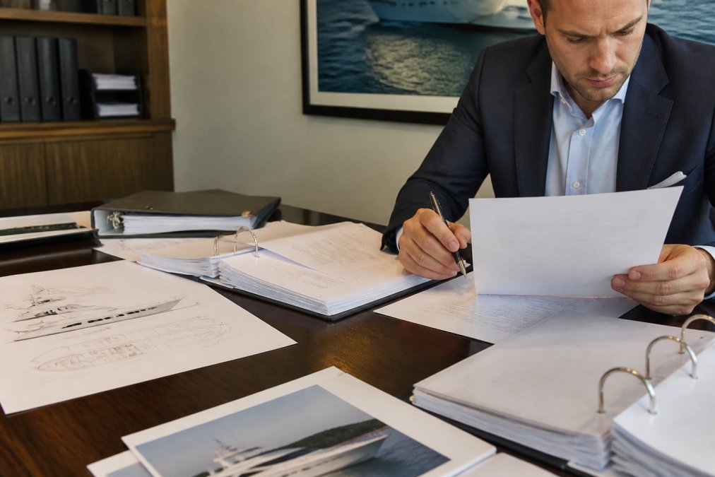 Professional reviewing vessel financing documentation and loan agreements at a modern office desk with yacht specifications and survey reports visible