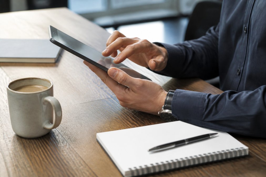 Close-up of hands using a tablet to complete online registration form, coffee cup and notepad visible on modern desk, natural office lighting