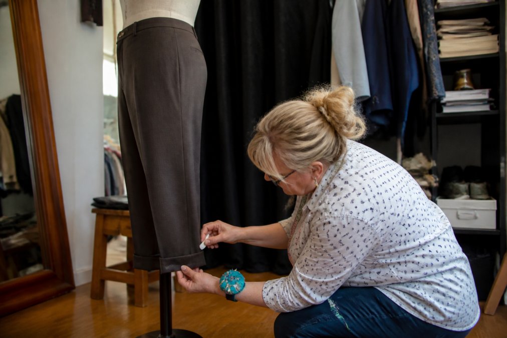 Professional seamstress carefully measuring and pinning the hem of tailored dress pants on a fitting form, using precision tools and marking the exact length with fabric chalk