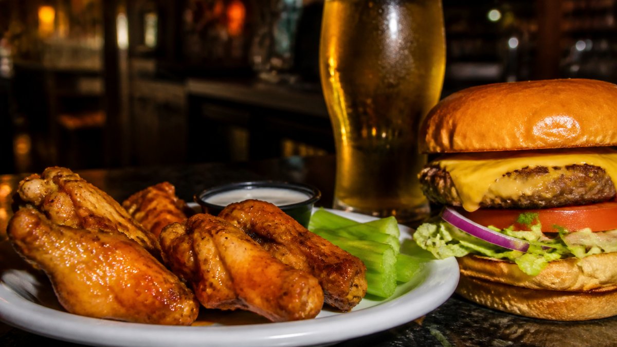 Close-up of a plate of crispy wings and a thick burger with cold beer on a dark bar counter, late evening lighting with warm amber bar lights in the background