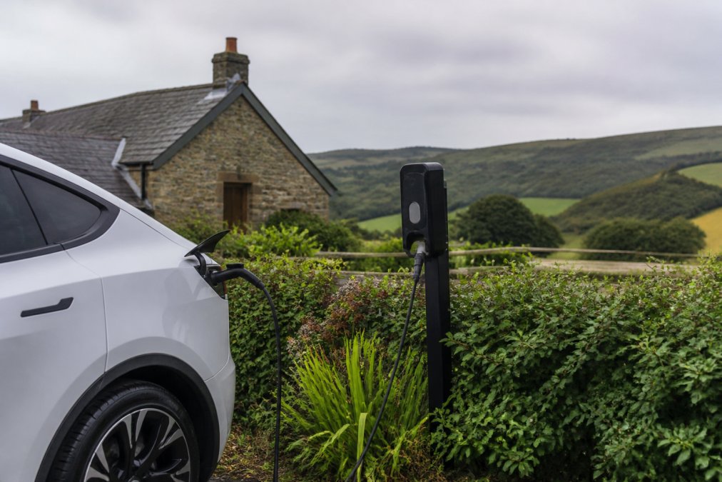 An electric vehicle parked at a charging station in a rural countryside setting, with a traditional stone holiday cottage visible in the background and rolling green hills under overcast sky