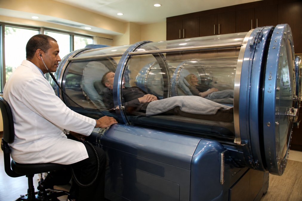 Board-certified hyperbaric physician monitoring vital signs while a patient receives treatment inside a multi-person clinical hyperbaric chamber in a modern medical office setting