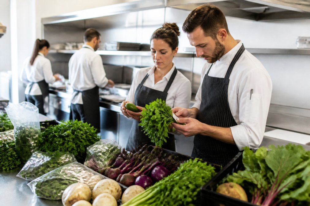 Professional catering staff inspecting fresh produce quality, examining vegetables and checking labels on packaging in a bright commercial kitchen setting with stainless steel benches