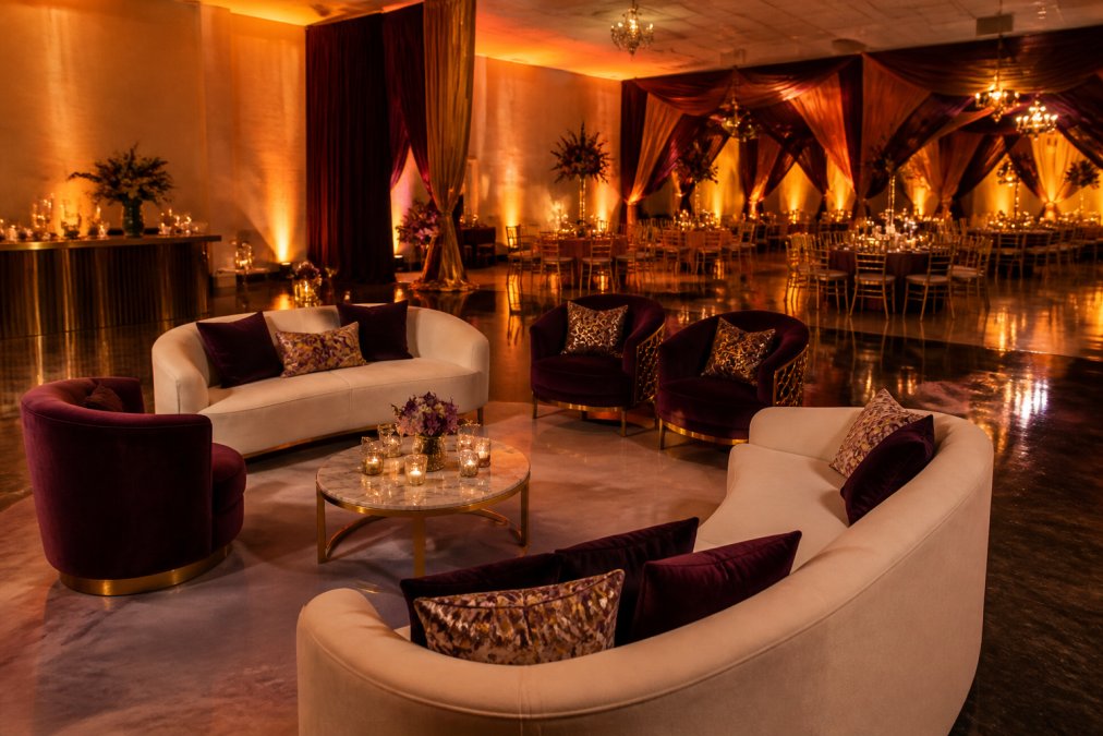 Elegantly decorated event space with dramatic amber and warm white up-lighting illuminating luxurious lounge furniture, sophisticated venue draping in jewel tones, and polished floors reflecting the ambient lighting creating an opulent ambiance for a formal celebration