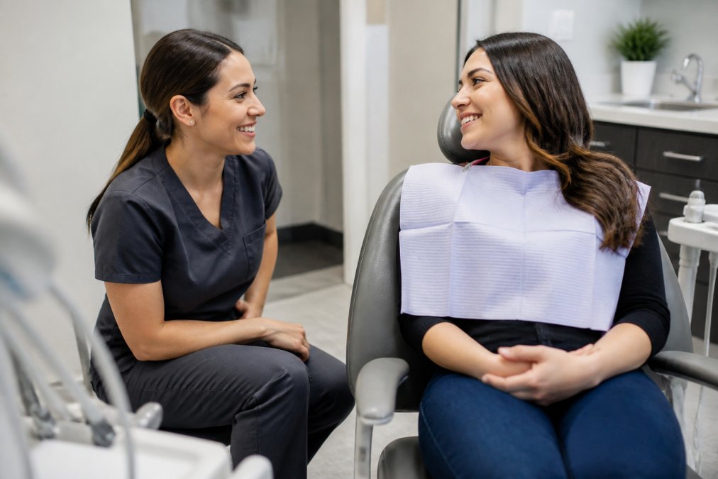 A dental hygienist kneeling beside a seated patient in a bright treatment room, both smiling during a relaxed conversation, with modern dental equipment visible in the background
