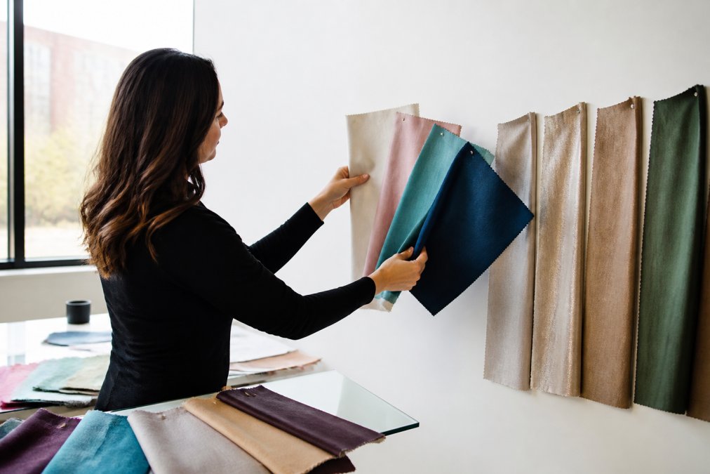 Event planner holding multiple fabric swatches in different colors and textures, examining them against a white wall in natural daylight, comparing how each shade appears under different lighting conditions