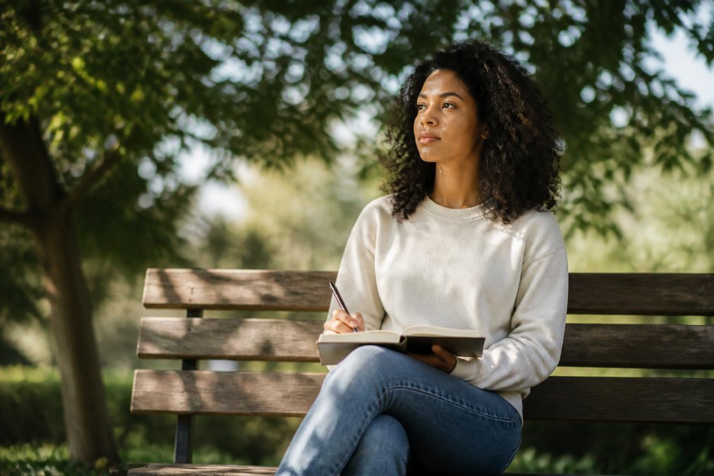 Person sitting on a wooden bench outdoors in dappled sunlight, journal open on lap, pen in hand, looking peaceful and contemplative with trees softly blurred in background