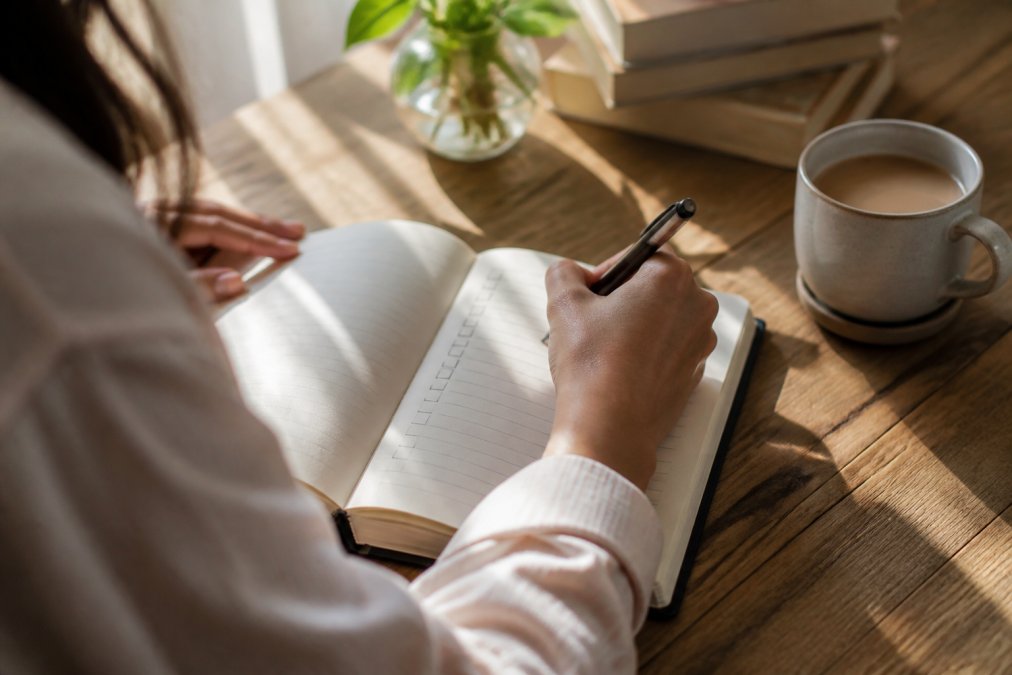 Close-up of hands holding a pen, writing a checklist in an open planner with warm coffee cup nearby on wooden desk, soft natural light streaming across the page