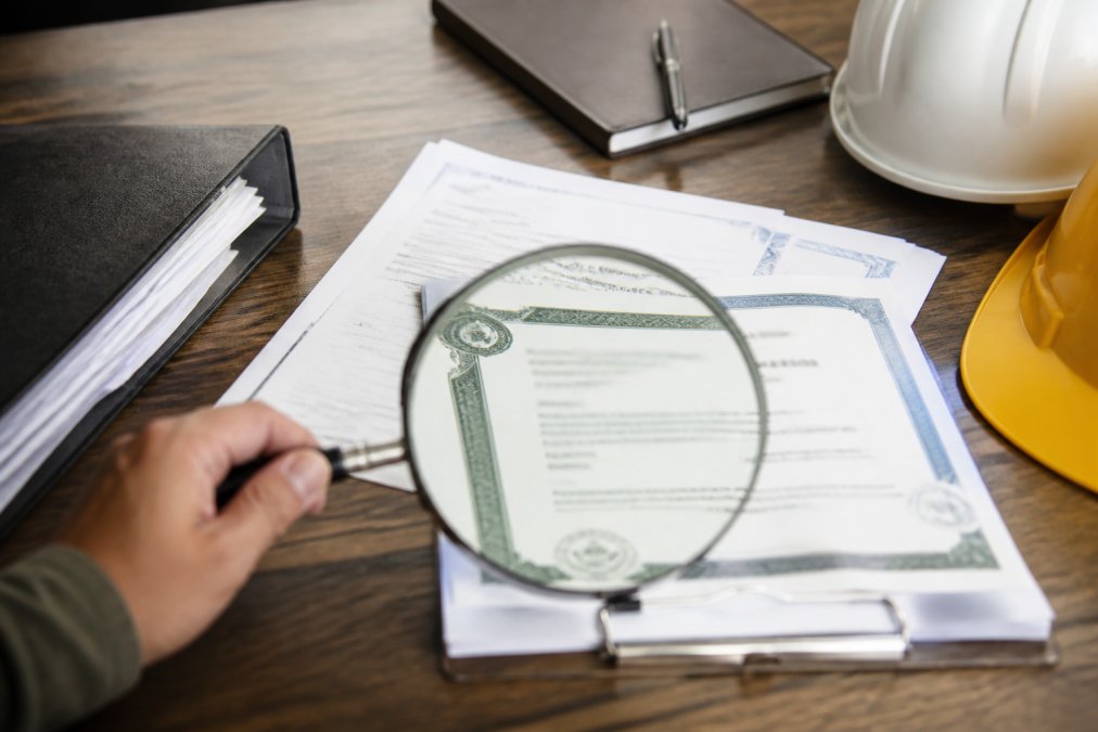 Close-up of professional roofing contractor's license documents, insurance certificates, and credentials arranged on a wooden desk with a magnifying glass examining the details