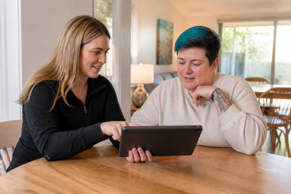 Support coordinator and NDIS participant reviewing specialist disability accommodation options together on a tablet in a comfortable home setting, discussing property features and accessibility requirements with natural afternoon lighting