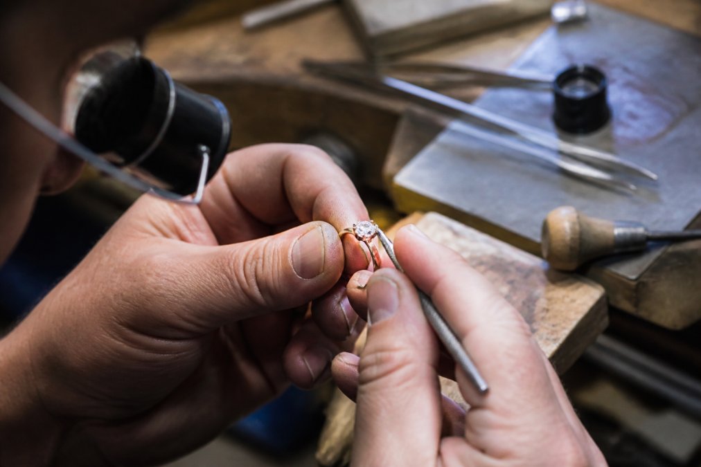 A jeweler's hands using precision tools to tighten a prong on a diamond engagement ring at a well-lit workbench, with loupe and tweezers nearby