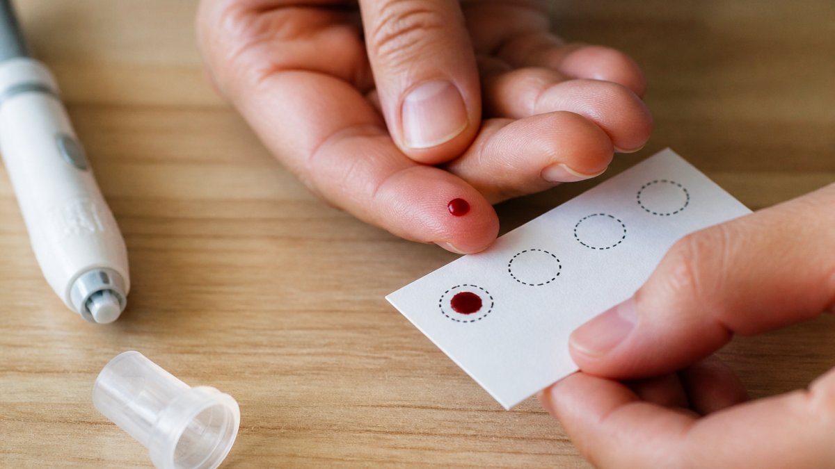 Close-up of a person's fingertip during finger-prick blood collection, showing a small drop of blood being collected onto a dried blood spot card with a lancet device visible, natural lighting