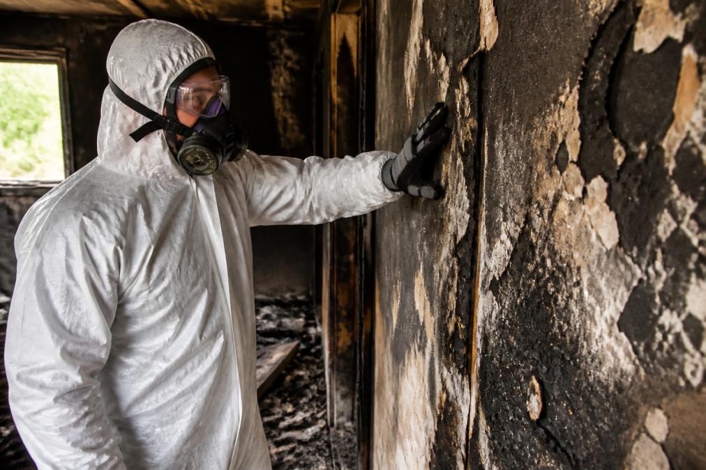 Restoration technician in full protective gear including respirator mask, safety glasses, and heavy-duty gloves carefully inspecting fire-damaged interior walls with visible soot and charred material buildup