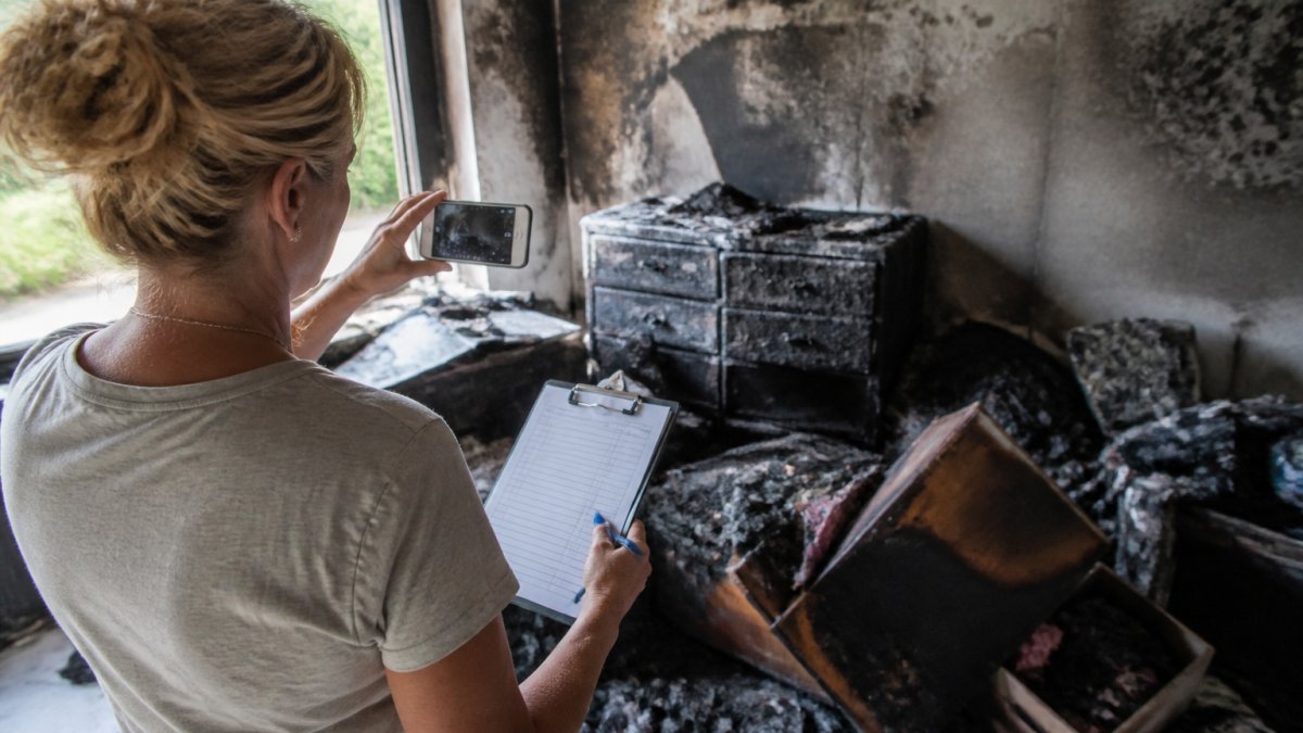 A homeowner in a fire-damaged room photographing charred belongings with a smartphone while holding a clipboard with inventory notes, smoke-stained walls visible in background
