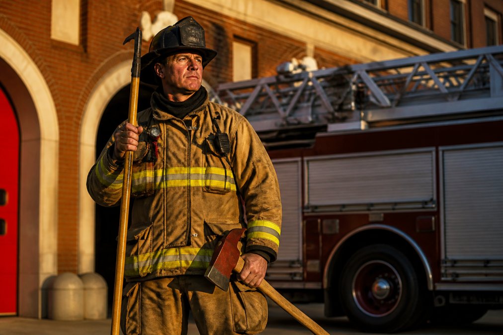 Firefighter in full turnout gear holding pike pole and fire axe standing in front of firehouse with ladder mounted on apparatus, golden afternoon light illuminating the scene