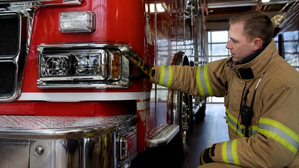 Firefighter in turnout gear inspecting front headlights and emergency lights on a red fire truck in a station bay