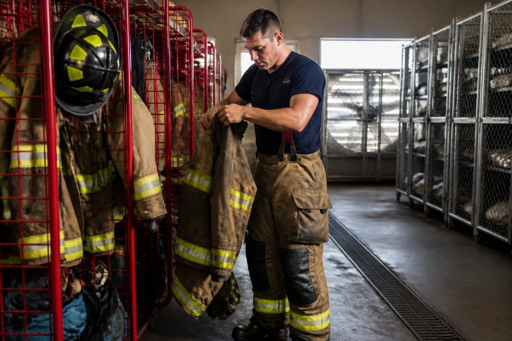 Firefighter in full turnout gear carefully removing and storing protective equipment in a dedicated gear storage area after a structure fire, demonstrating proper contamination-aware handling with organized gear racks and ventilation