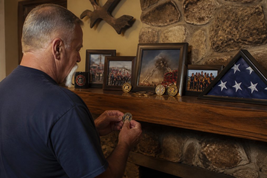 A retired firefighter displaying commemorative challenge coins and engraved keepsakes arranged on a wooden fireplace mantle, with framed photos of his fire service career and a folded American flag nearby, warm evening light