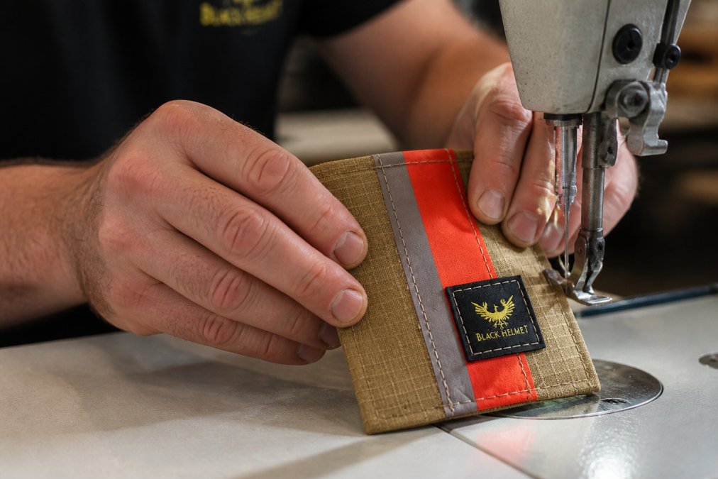 Close-up of artisan hands carefully crafting a wallet from authentic decommissioned fire service bunker gear material, showing detailed stitching and texture of worn equipment, indoor workshop lighting with natural window light