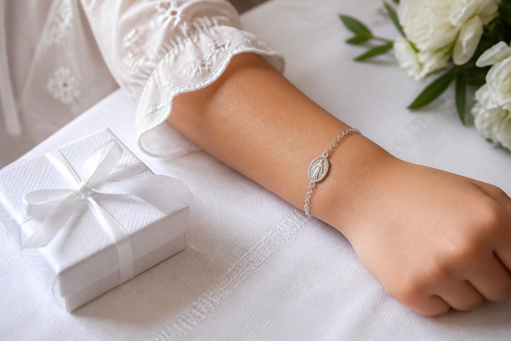A close-up of a young girl's wrist wearing a delicate silver Miraculous Medal bracelet, with a soft white dress sleeve visible, resting on a white linen tablecloth next to a small gift box