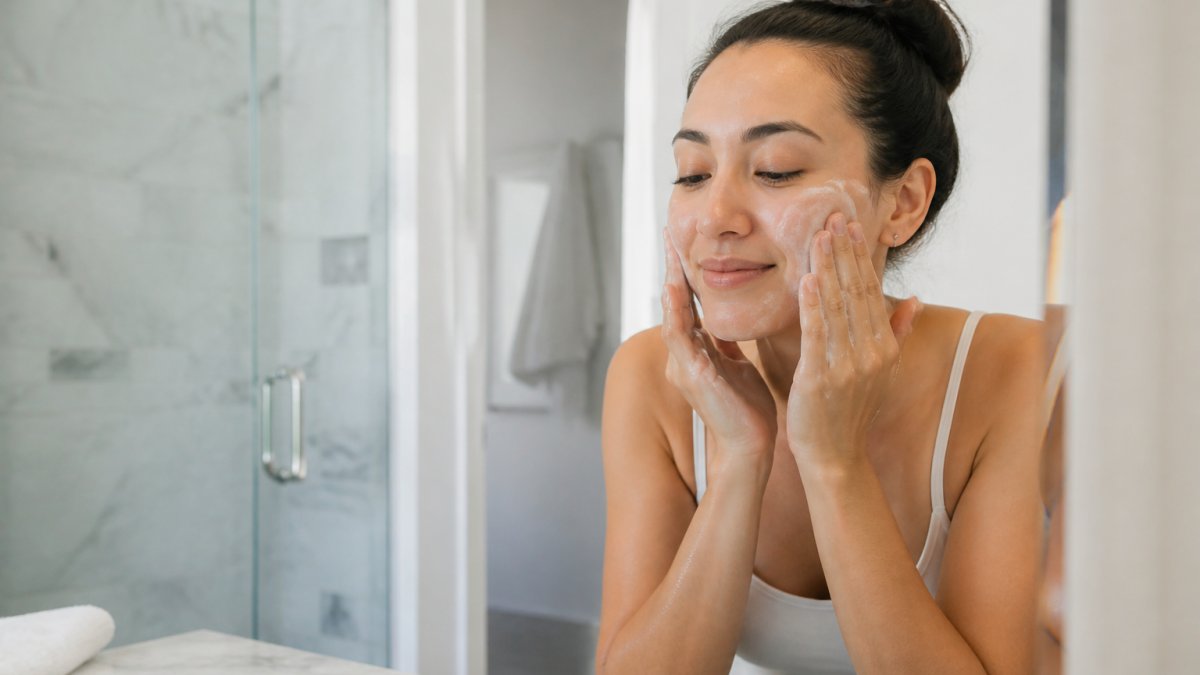 Close-up of a woman with clean, makeup-free eyes gently washing her face with a soft cleanser in a bright bathroom, preparing for a lash appointment
