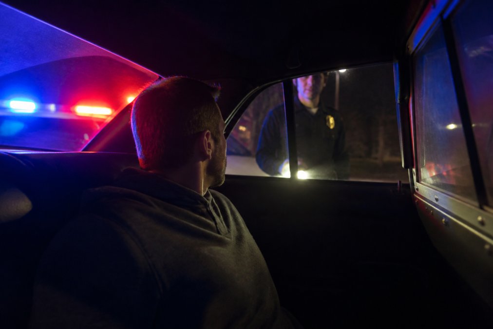 Person sitting in back of police car during traffic stop at night, officer visible through window, red and blue lights illuminating the scene