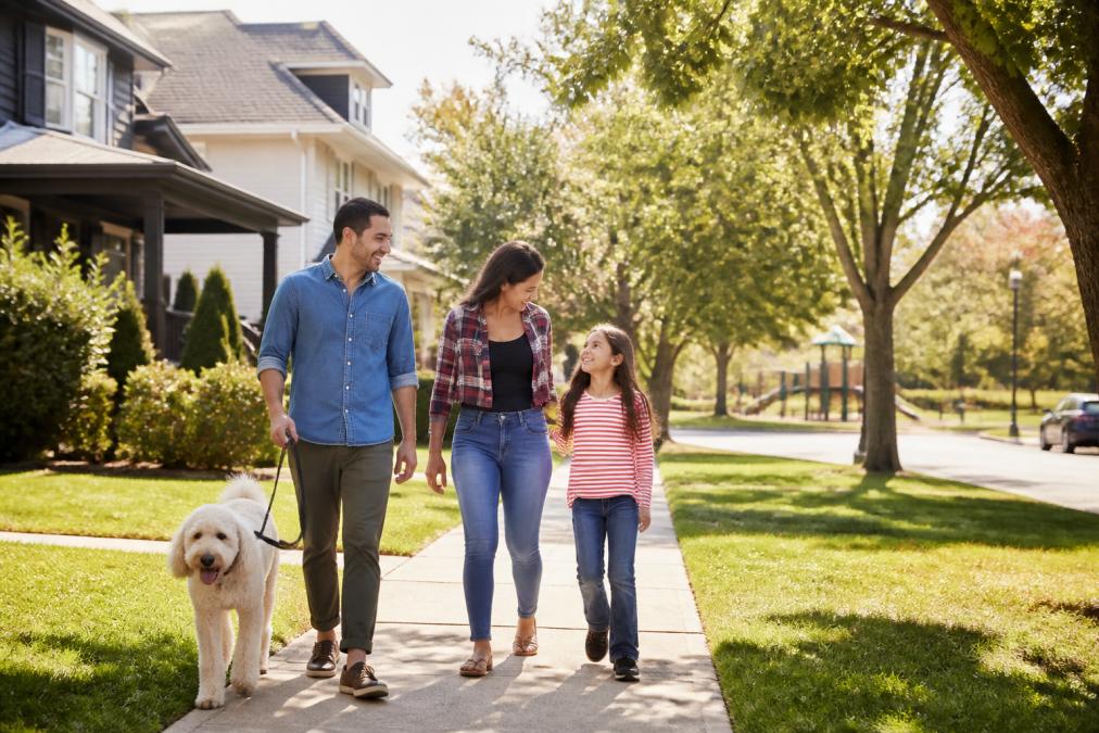 Family walking through a tree-lined residential North Dallas street with modern homes, manicured lawns, and a community park visible in the distance on a clear, sunny afternoon