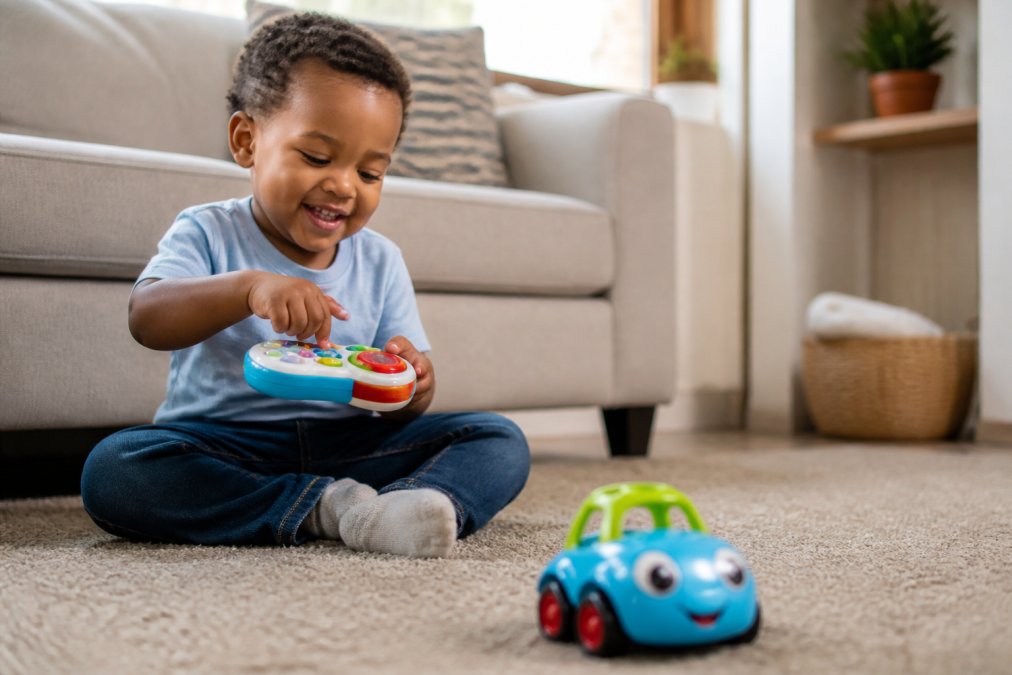 A young child sitting on a living room floor, smiling and pressing buttons on a colourful toy remote control, with a small toy car nearby