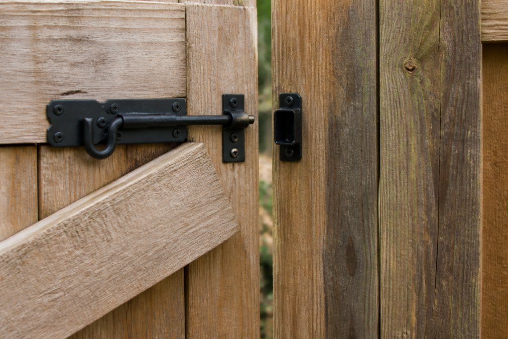 Close-up of a wooden gate latch misaligned with the strike plate on a fence post, showing the gap that prevents it from closing