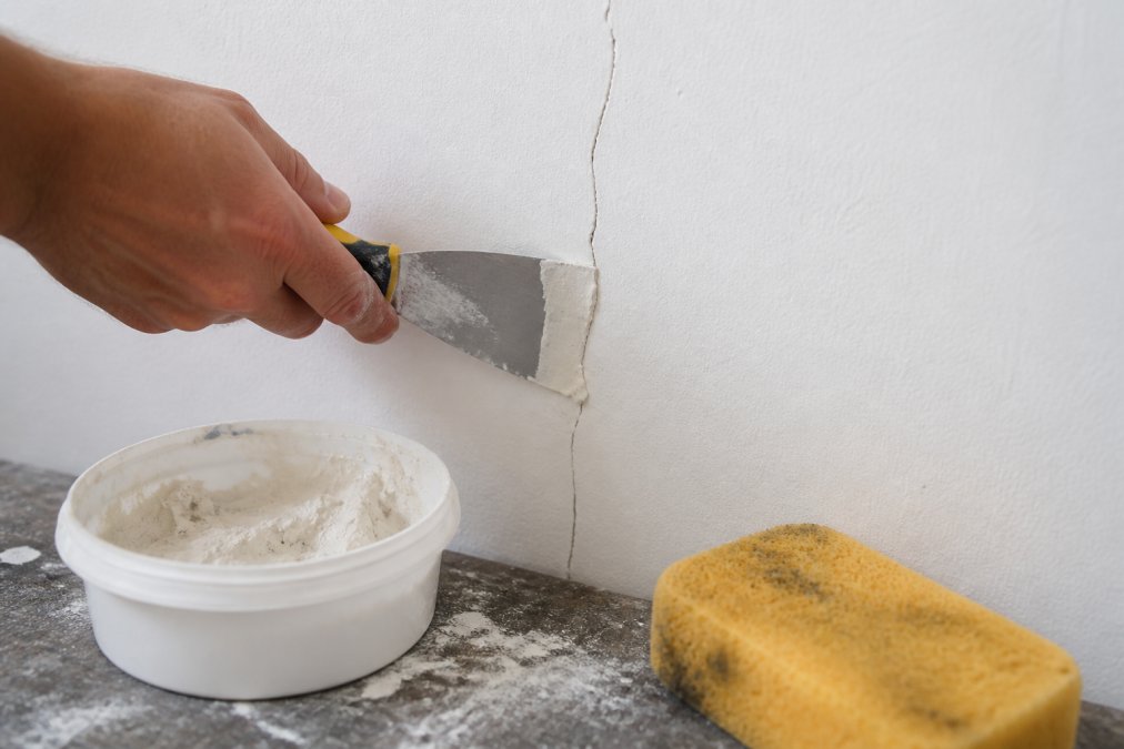 Close-up of a filling knife applying joint compound into a hairline crack in white plaster wall, with a damp sponge and container of filler visible on a work surface