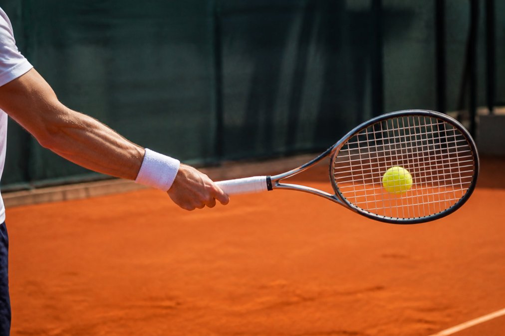 Close-up of a player's arm and racquet at contact point with the ball, showing a slightly closed racquet face angle with the player's wrist in neutral position, photographed from the side during afternoon sunlight on a clay court