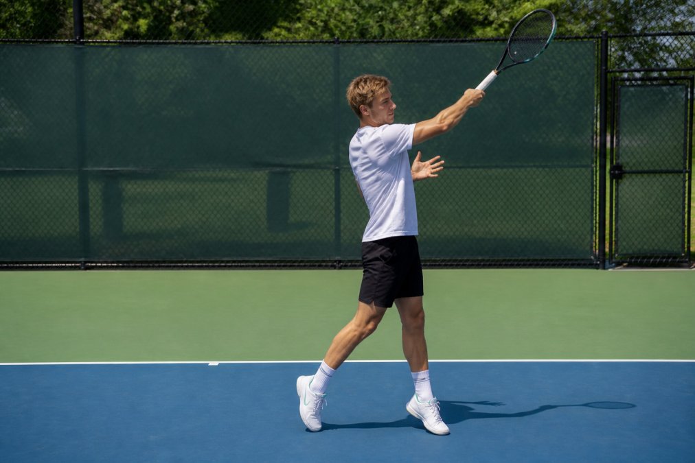 Player demonstrating the low-to-high brushing motion during a forehand stroke, showing the vertical swing plane from the side angle, with the racquet starting below the waist and finishing above the shoulder on a bright outdoor court