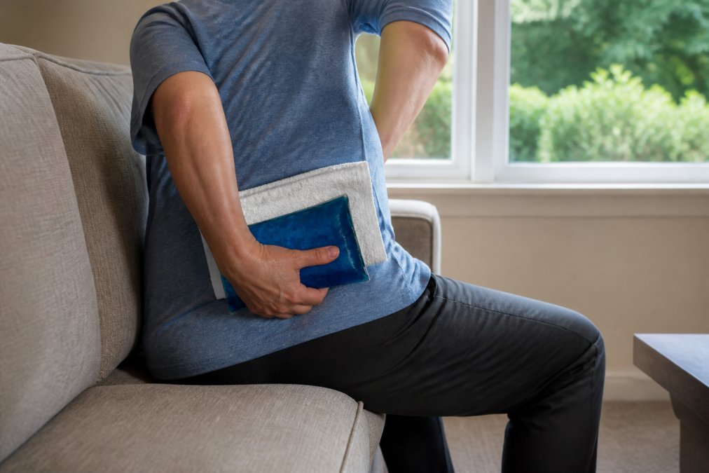 Person applying ice pack to lower back while seated on couch, showing proper ice application technique with towel barrier between skin and pack