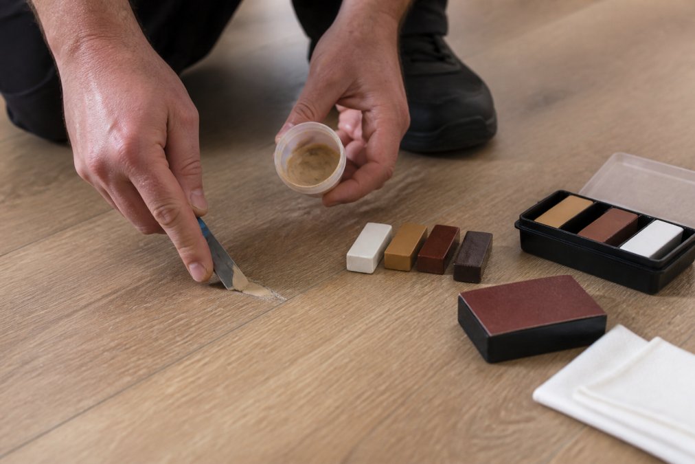 Hands carefully applying filler compound to a small chip in laminate flooring using a specialist tool, with repair materials, sandpaper, and finishing supplies arranged on the surface nearby