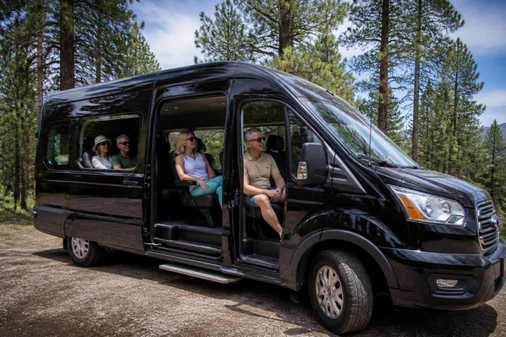 A comfortable Ford Transit van with passengers visible through large windows, scenic pine forests and high-elevation Arizona landscape visible outside, natural daylight illuminating the interior