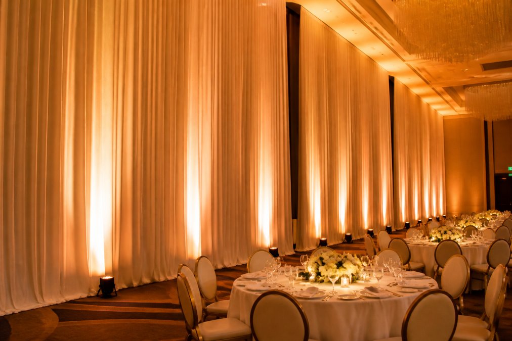 Elegant hotel ballroom set up for a corporate gala with tall white pipe and drape panels lining the walls at approximately 20 feet high, soft amber uplighting casting warm color on the fabric, round banquet tables with white linens in the foreground