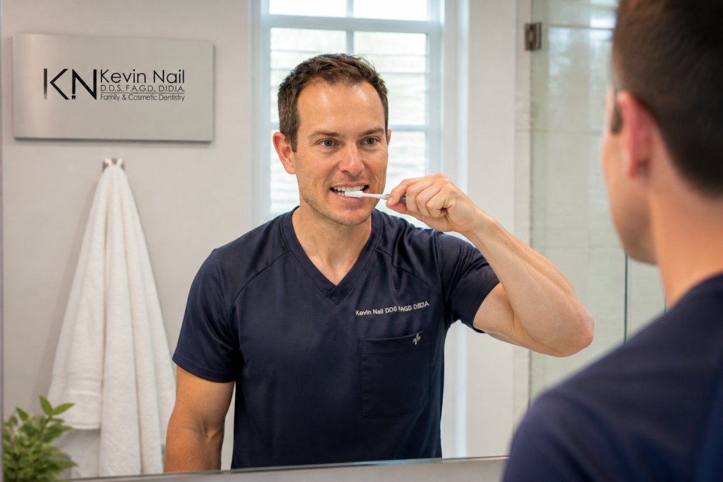 Patient carefully brushing teeth with soft-bristled toothbrush near implant site, demonstrating gentle oral hygiene technique with proper lighting in home bathroom setting