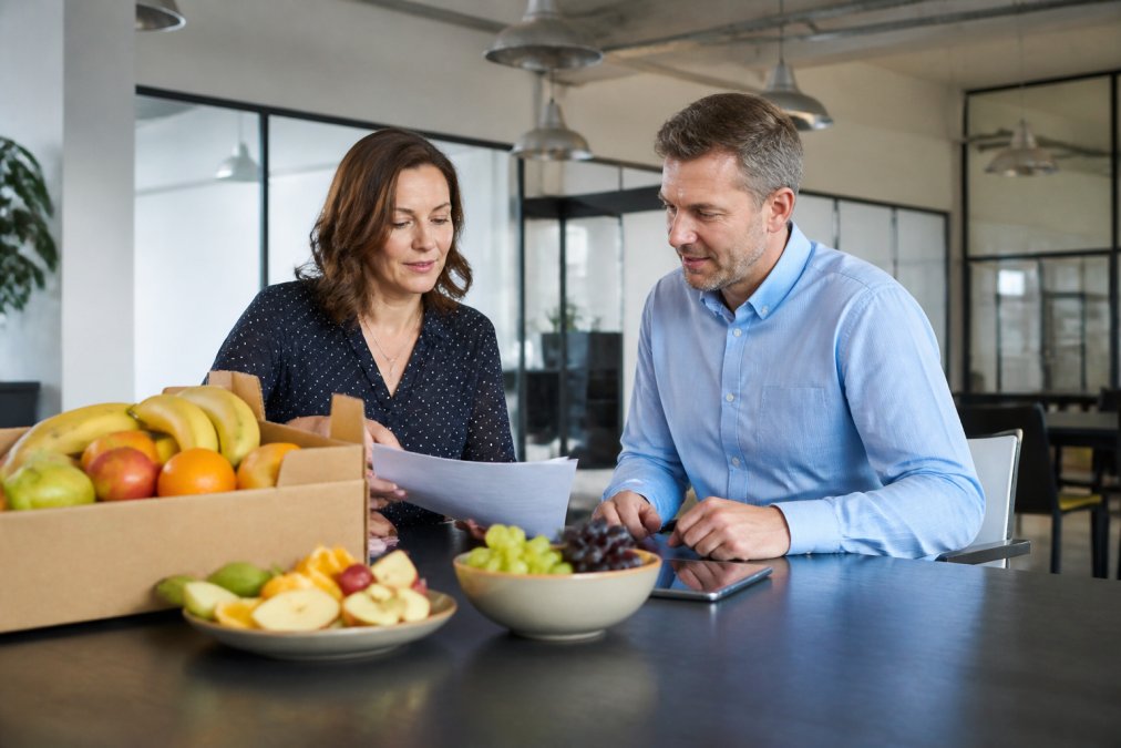 Kontorchef og indkøbsansvarlig gennemgår frugtordning på computer med tablet på skrivebordet, frugtkasse og frugtprøver synlige