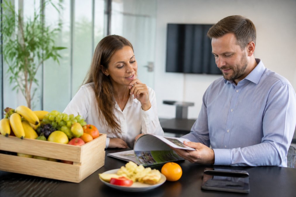 Kontorchef og indkøbsansvarlig planlægger fleksibel frugtlevering ved computer med frugtkasse og prøver på skrivebordet.