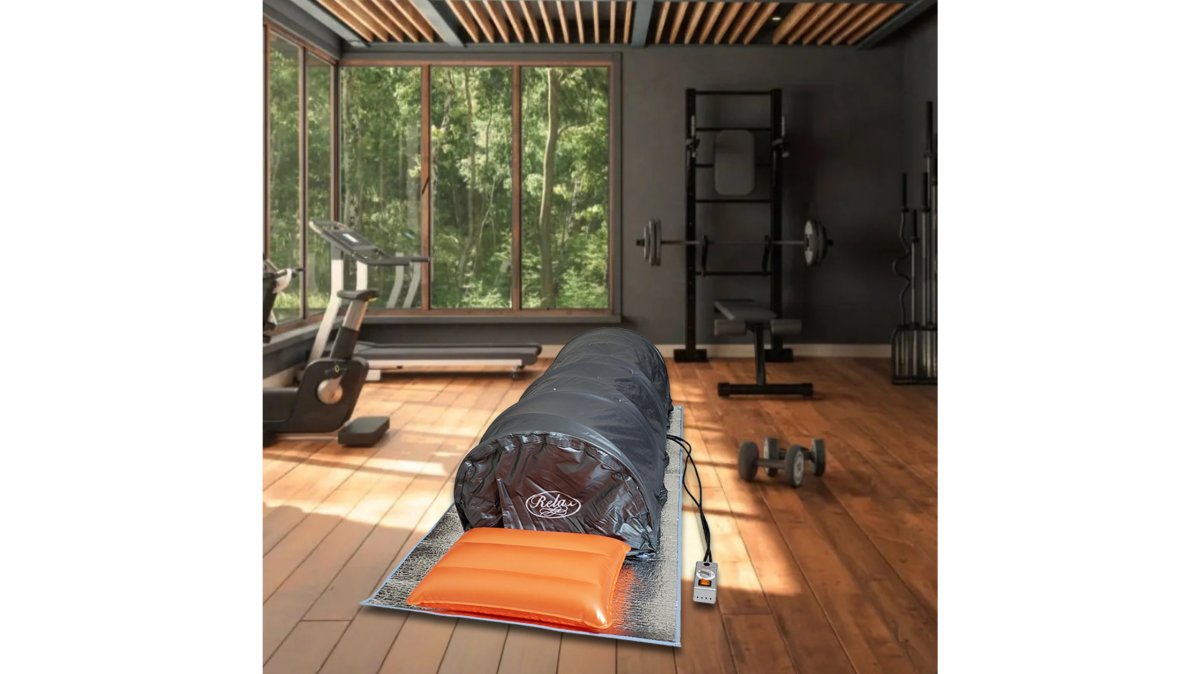 A person lying down comfortably inside a portable infrared sauna tent, zipped up to the neck, with a calm expression and a water bottle nearby in a bright home living space