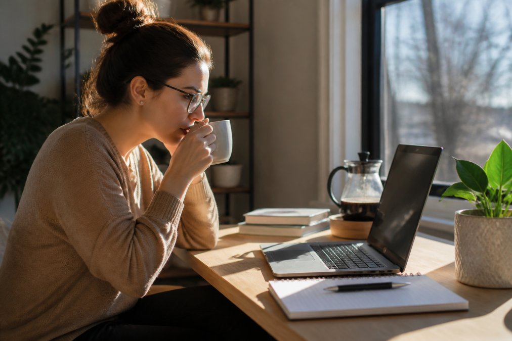 Remote worker seated at ergonomic home office desk with steaming coffee cup beside laptop, morning sunlight streaming through window onto workspace, focused expression