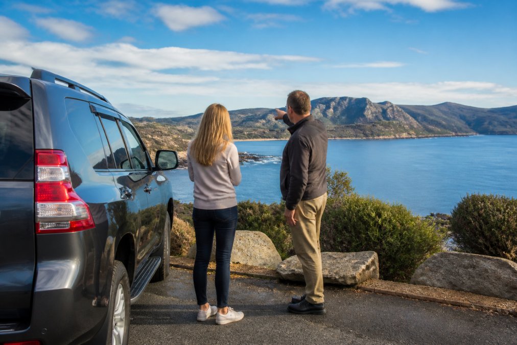 Solo traveler and guide standing by an SUV on a coastal road during flexible day tours of Hobart.