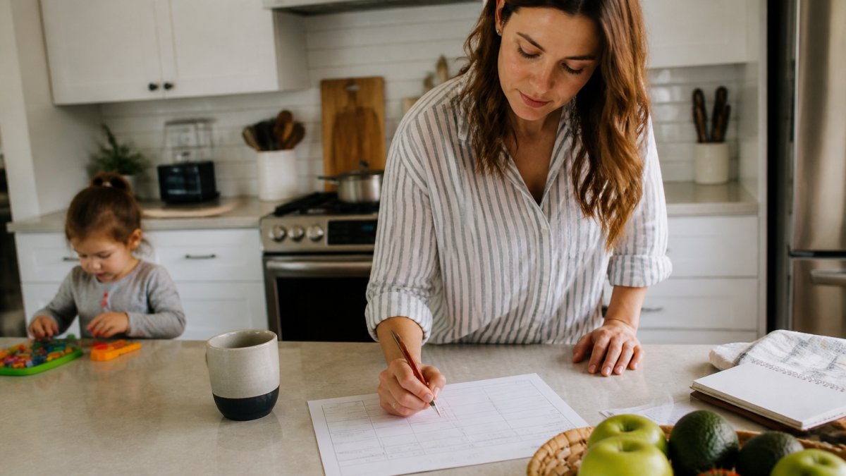 A parent organizing flexible meal plans on a notepad with a digital grocery list nearby in a busy kitchen.