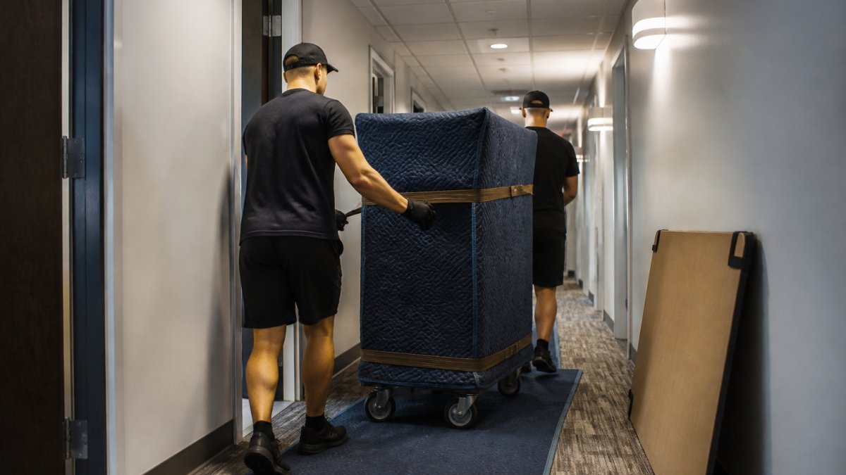 Professional crew carefully maneuvering large office furniture and equipment through narrow commercial hallway using protective padding and proper lifting equipment during evening hours