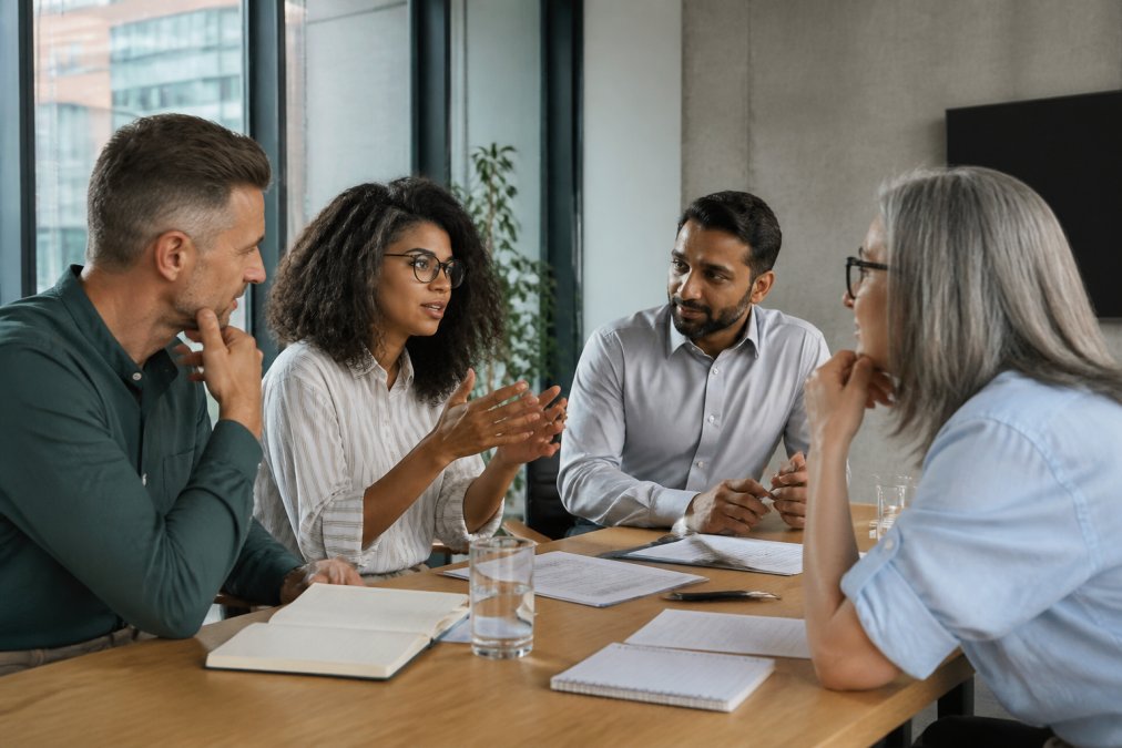 Diverse team of managers in modern office environment gathered around table in discussion, collaborative atmosphere, natural daylight, applying concepts in real conversation