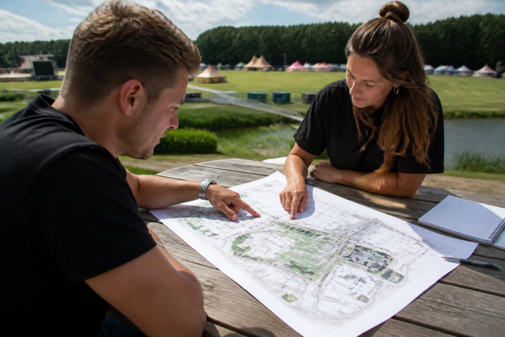 Eventplanner en klant bespreken outdoor reclame-opstellingen, wijzend naar verschillende locaties op een hoogteplattegrond van een evenemententerrein, met notities en maatstaven zichtbaar