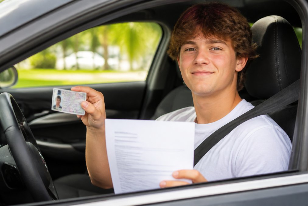 Young new driver sitting in driver's seat of car, holding driver's license and insurance documents, looking confident and ready