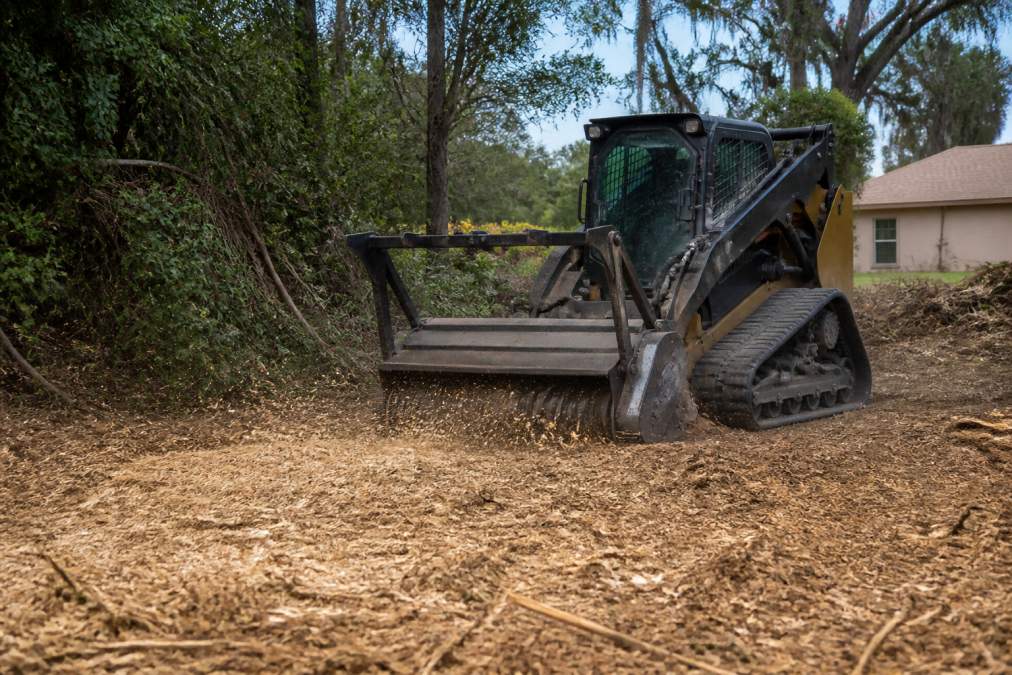 Heavy machinery with mulching attachment grinding trees and brush into fine mulch on a residential property, showing the immediate on-site transformation with wood chips covering the cleared ground