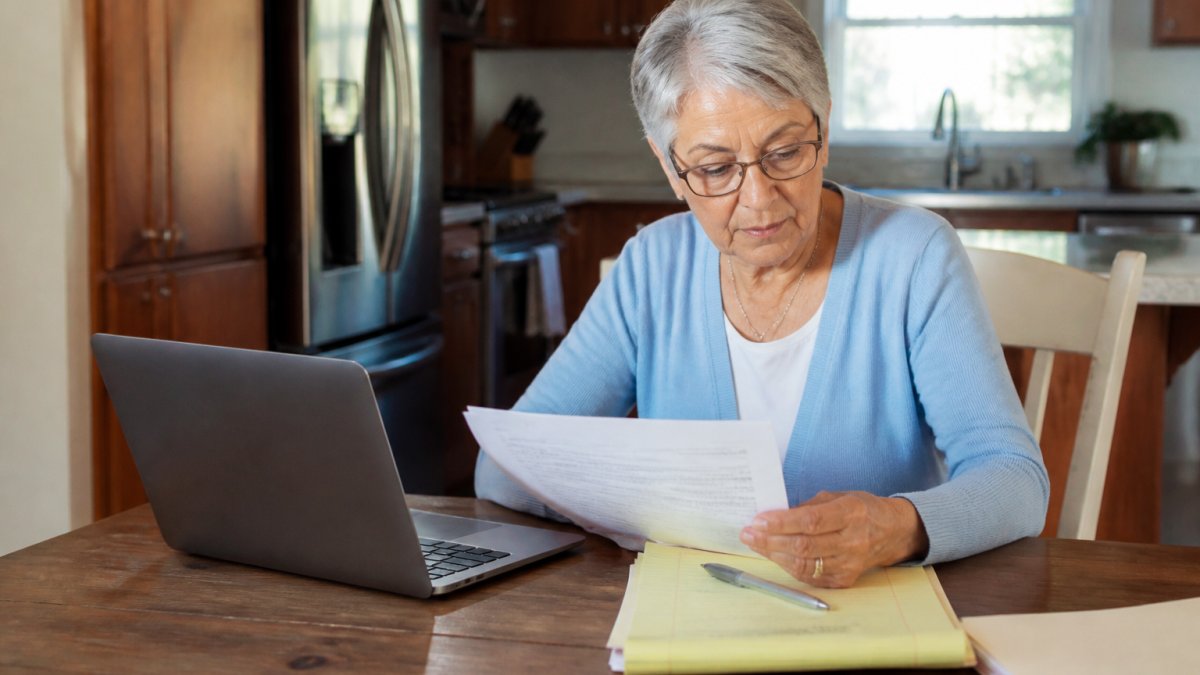 Older adult sitting at a kitchen table with a laptop and documents, reviewing Medicare plan options with a notepad and pen nearby, appearing focused and engaged