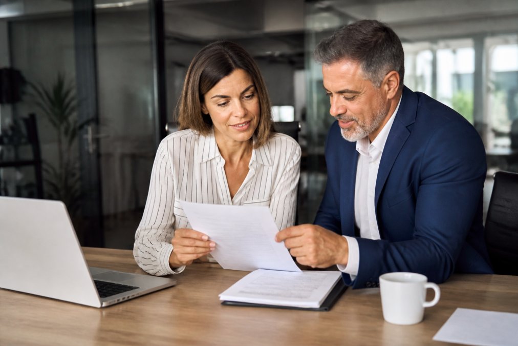 Business owner and attorney reviewing a contract document together at a desk with laptop and coffee, natural office lighting